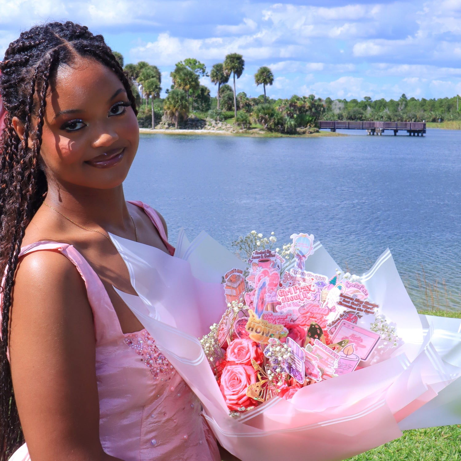 Young girl in a pink dress holding a bouquet of flowers by a lake with trees in the background.
